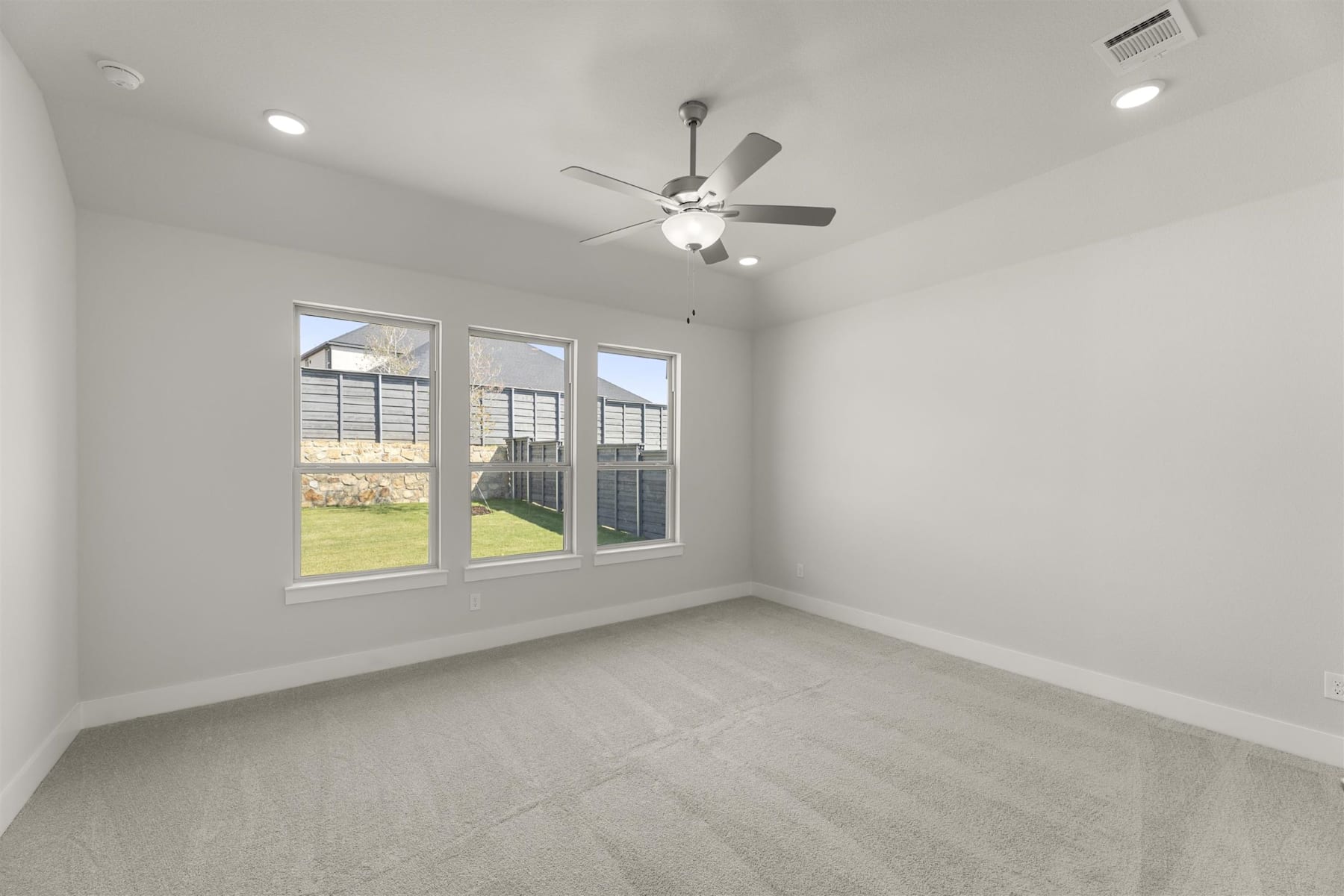 A spacious, well-lit room with a ceiling fan, large windows overlooking a grassy outdoor area, and a clean, minimalist interior design.