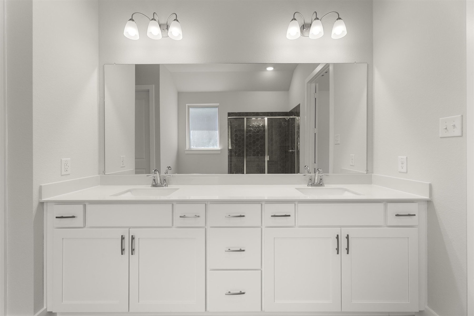 A modern, minimalist bathroom with a large vanity, double sinks, and overhead lighting fixtures against a white wall backdrop.