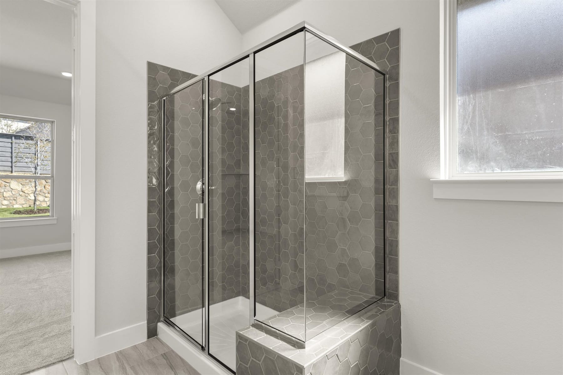 A modern, glass-enclosed shower enclosure with gray tile walls stands in a bright, airy bathroom setting.