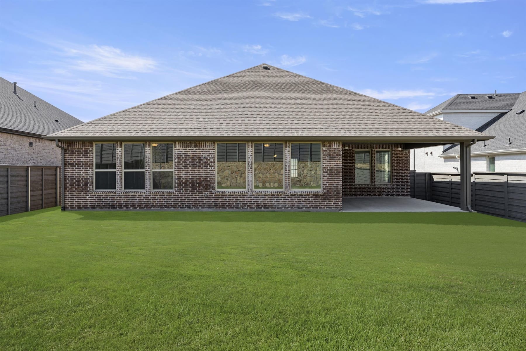 A single-story brick house with a slanted roof sits on a well-manicured lawn, surrounded by a clear blue sky with scattered clouds.