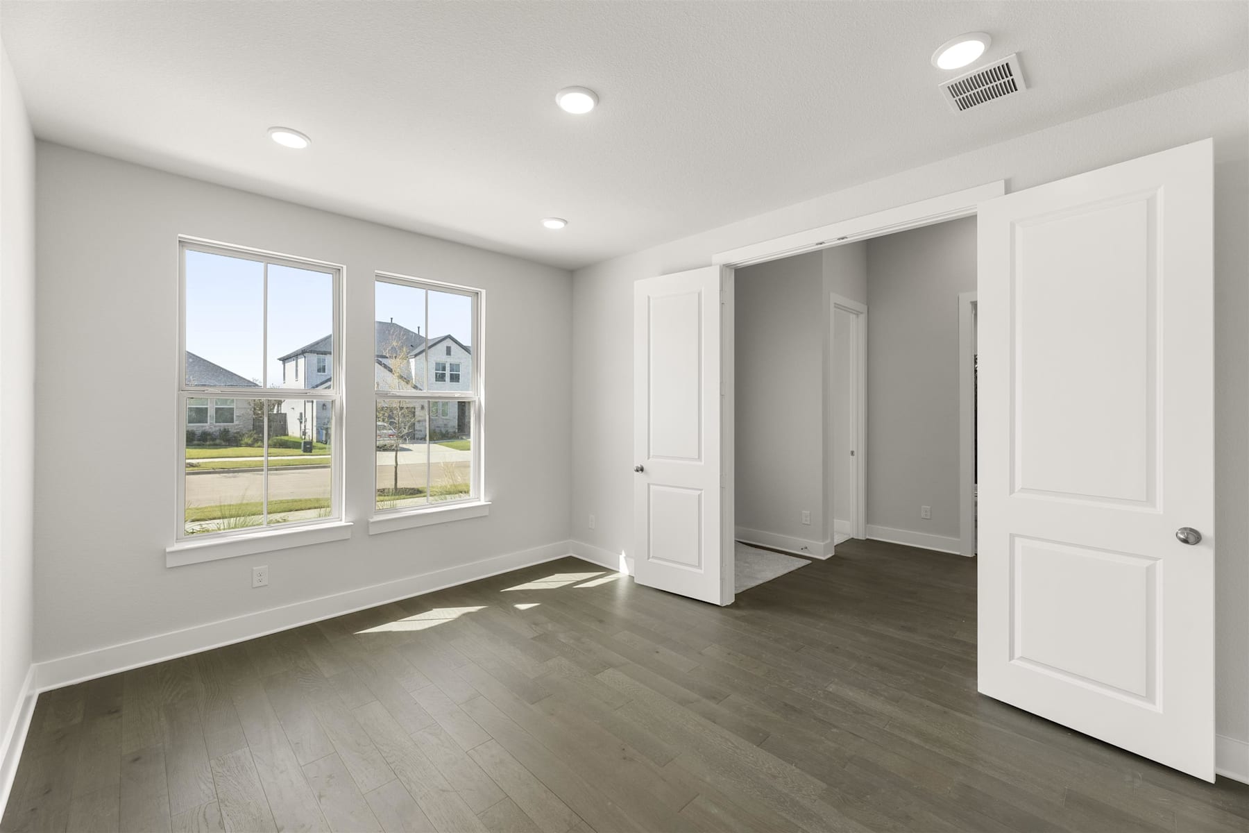 A spacious, well-lit room with white walls, dark hardwood floors, and a window overlooking a grassy outdoor area.