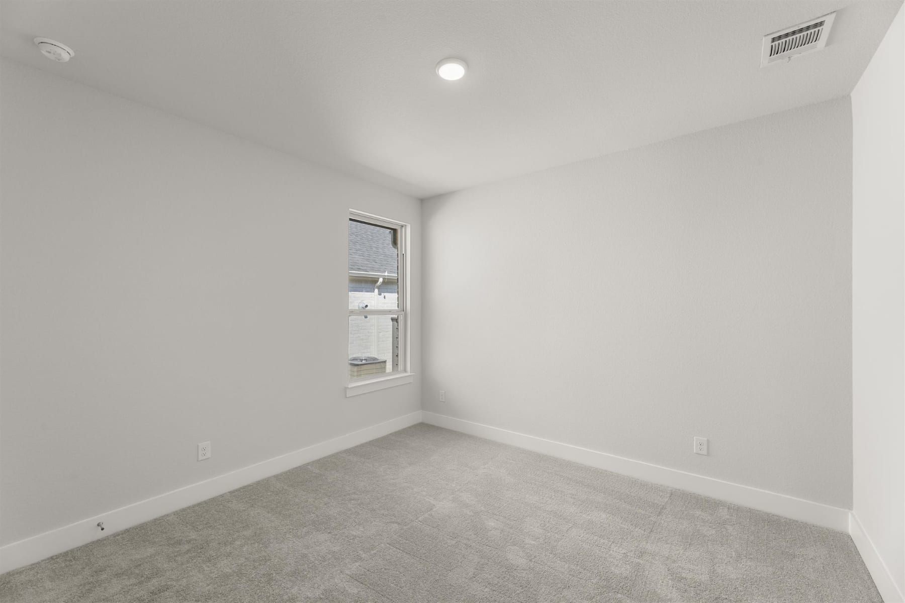 A simple, minimalist bedroom with a window, white walls, and gray carpeted floor.