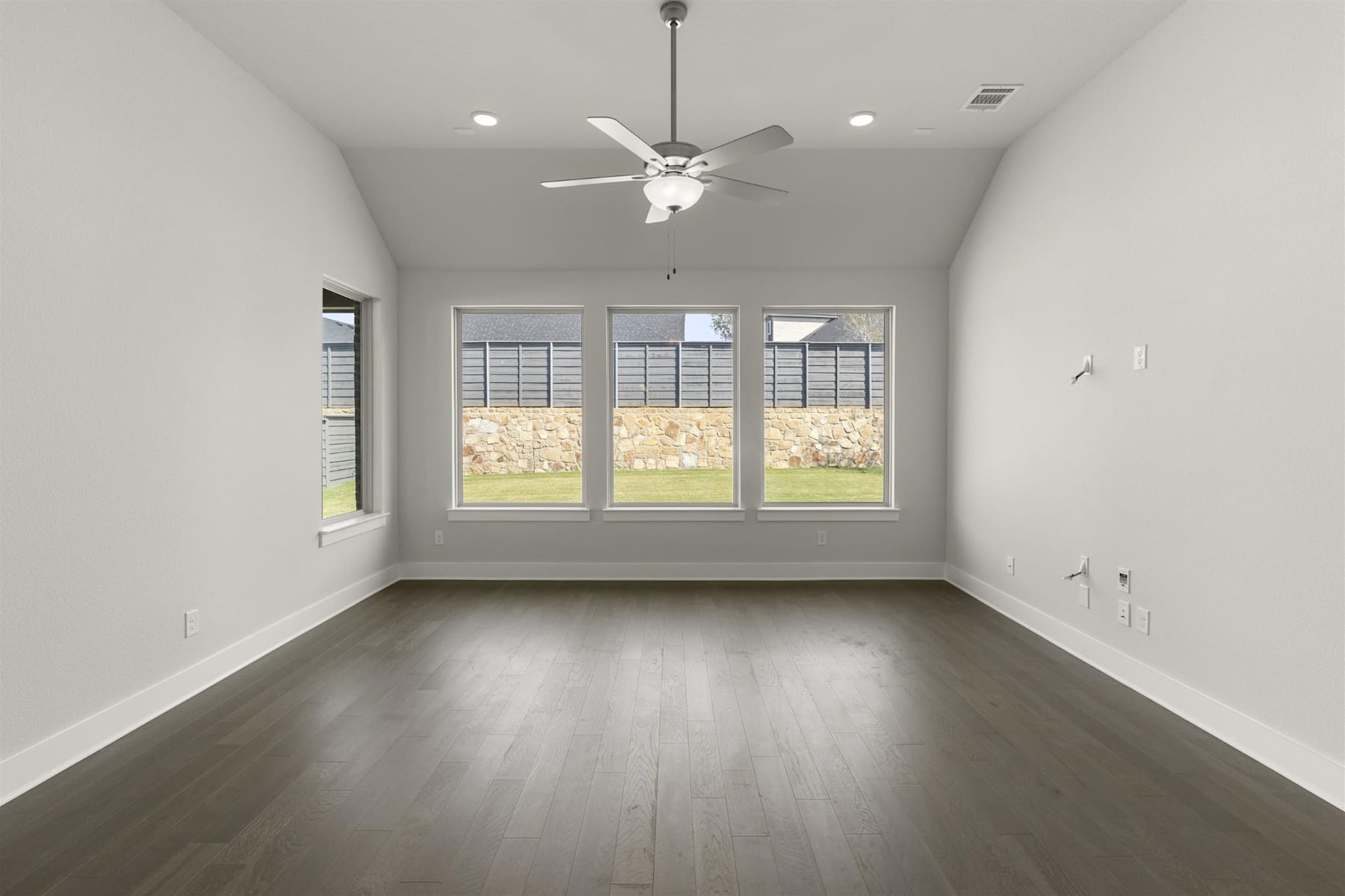 A spacious, well-lit room with a ceiling fan and large windows overlooking a backyard.