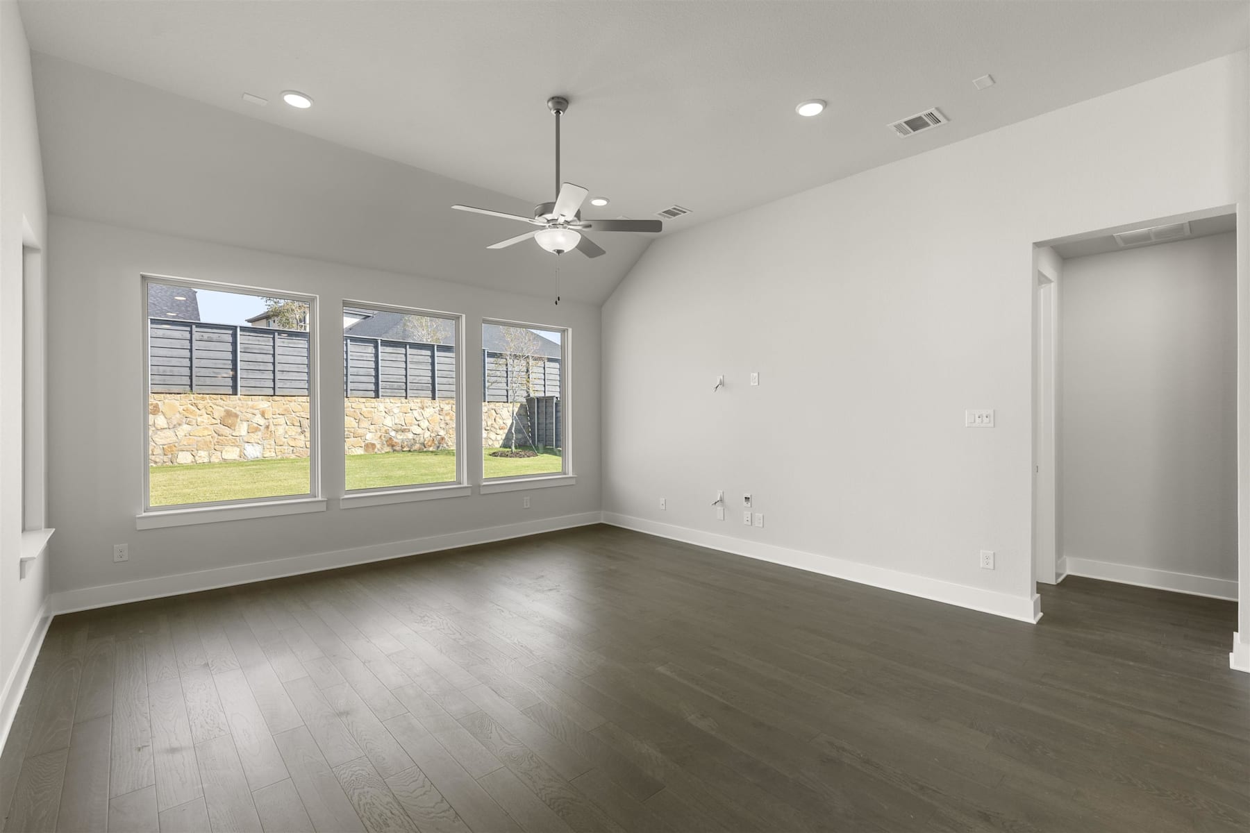 A spacious, well-lit room with a ceiling fan, large windows overlooking a yard, and dark hardwood floors.