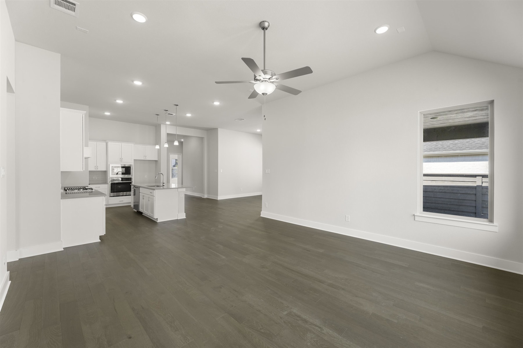 A spacious, open-concept living area with a kitchen visible in the background, featuring hardwood floors, a ceiling fan, and a large window providing natural light.