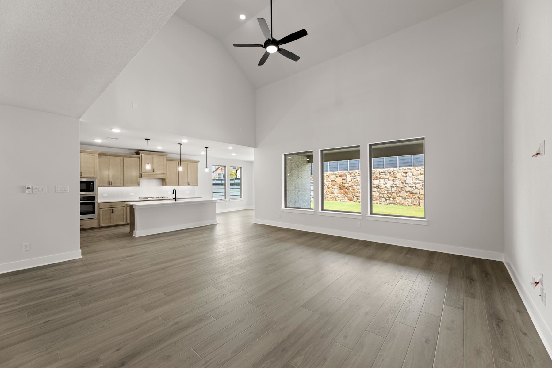 A spacious, modern living room with a high ceiling, hardwood floors, and a kitchen visible in the background. The room features large windows, allowing natural light to flood the space.