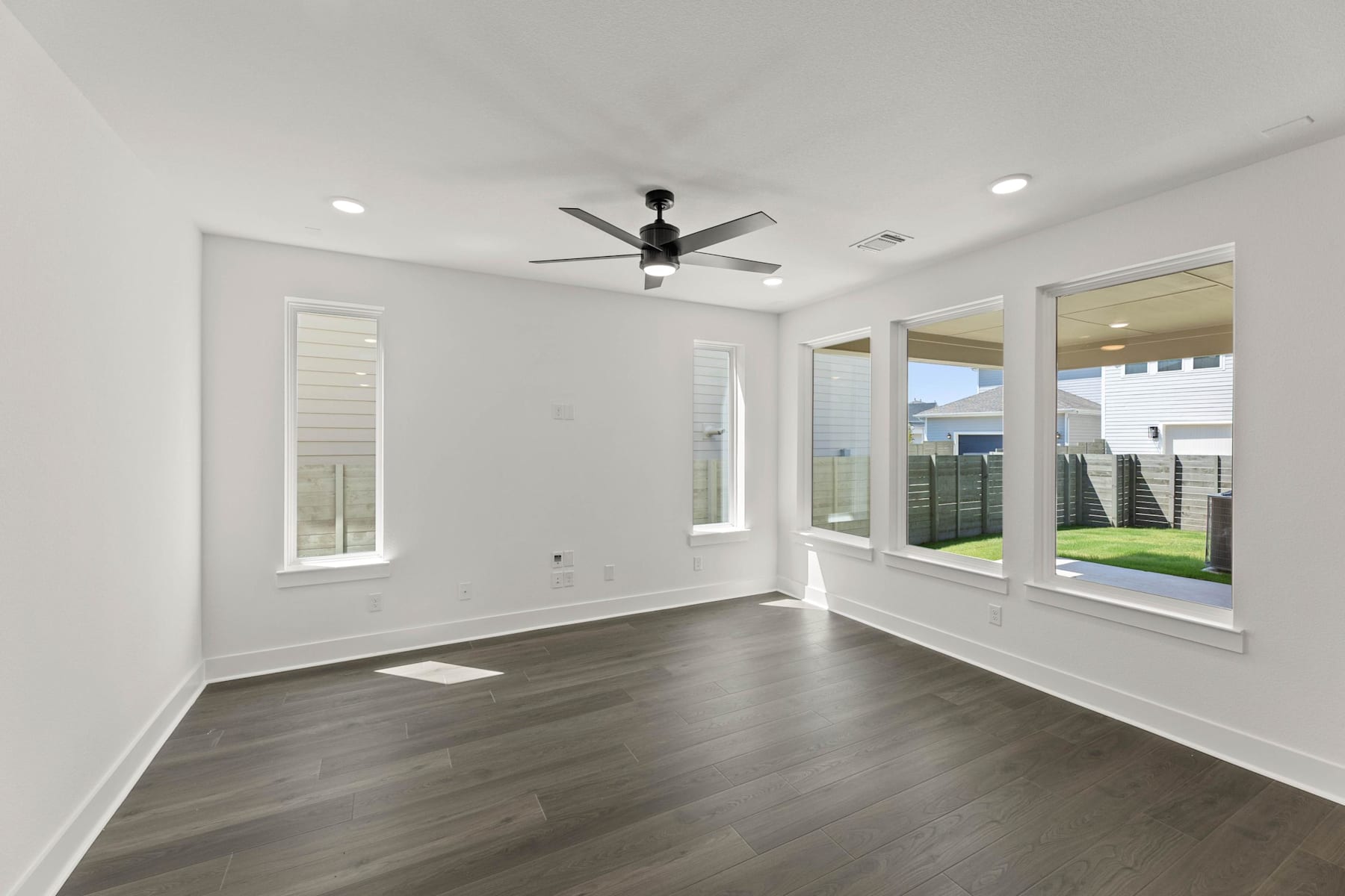 A spacious, well-lit room with white walls, dark hardwood floors, and a ceiling fan, overlooking a backyard through large windows.