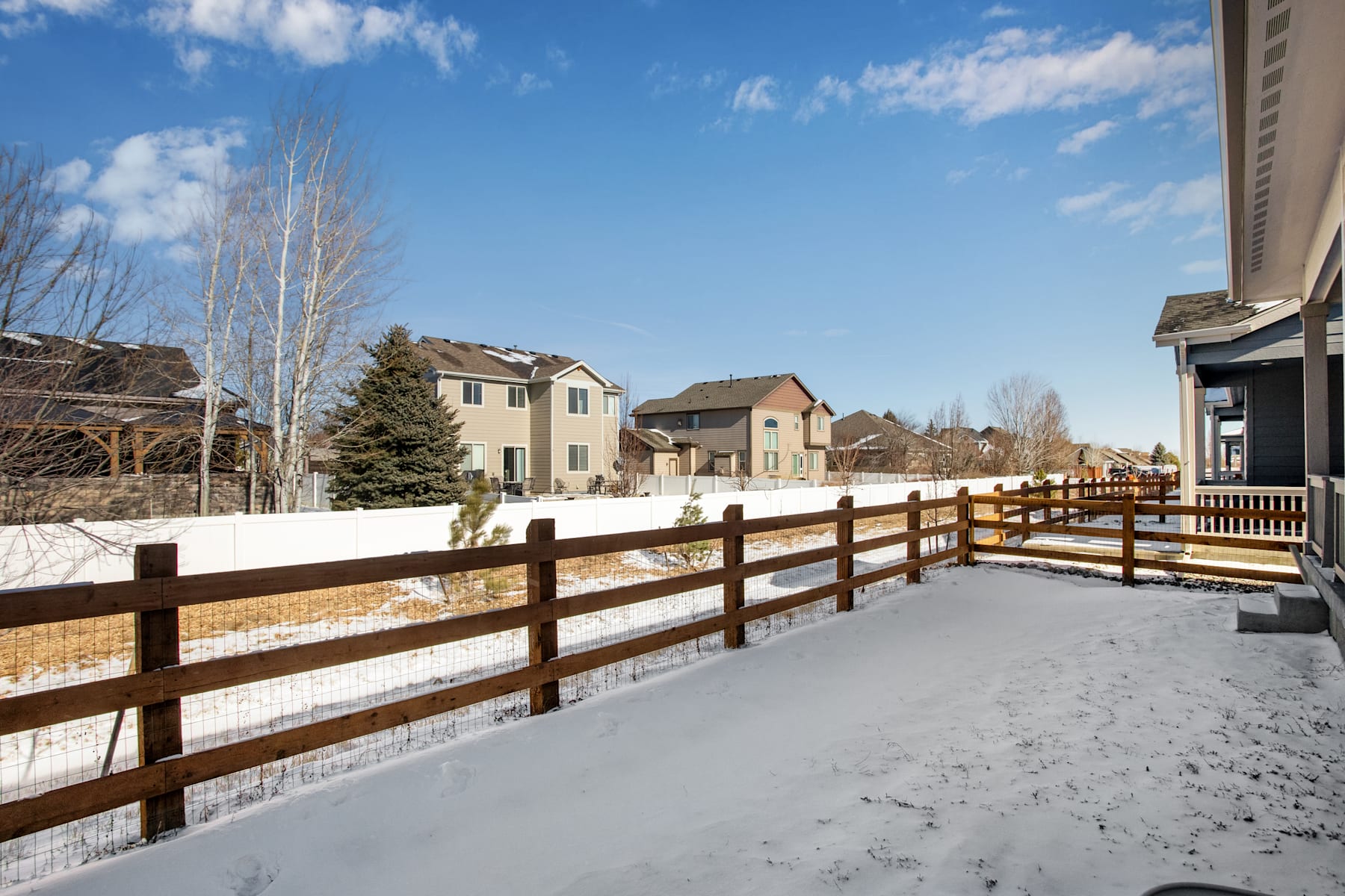 A snowy wooden fence runs along a path, with residential buildings visible in the background under a blue sky with scattered clouds.
