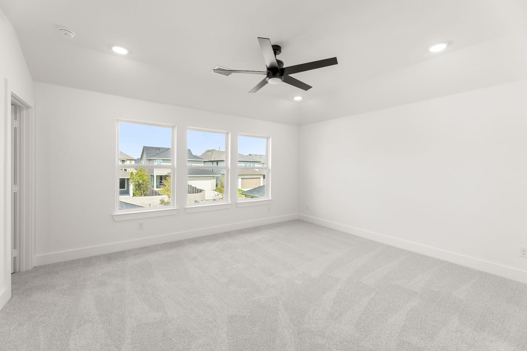 A spacious, well-lit room with a ceiling fan and large windows overlooking a residential neighborhood.