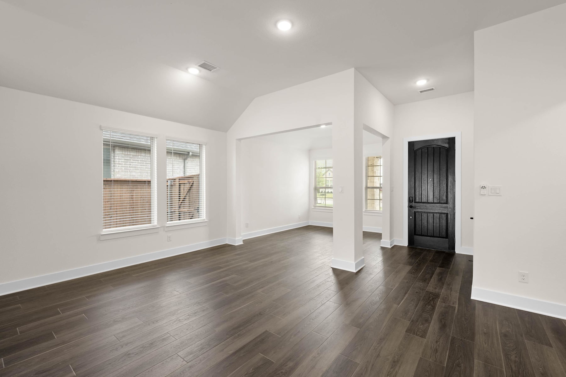 A spacious, well-lit room with white walls, dark hardwood floors, and a black wooden door visible in the background.