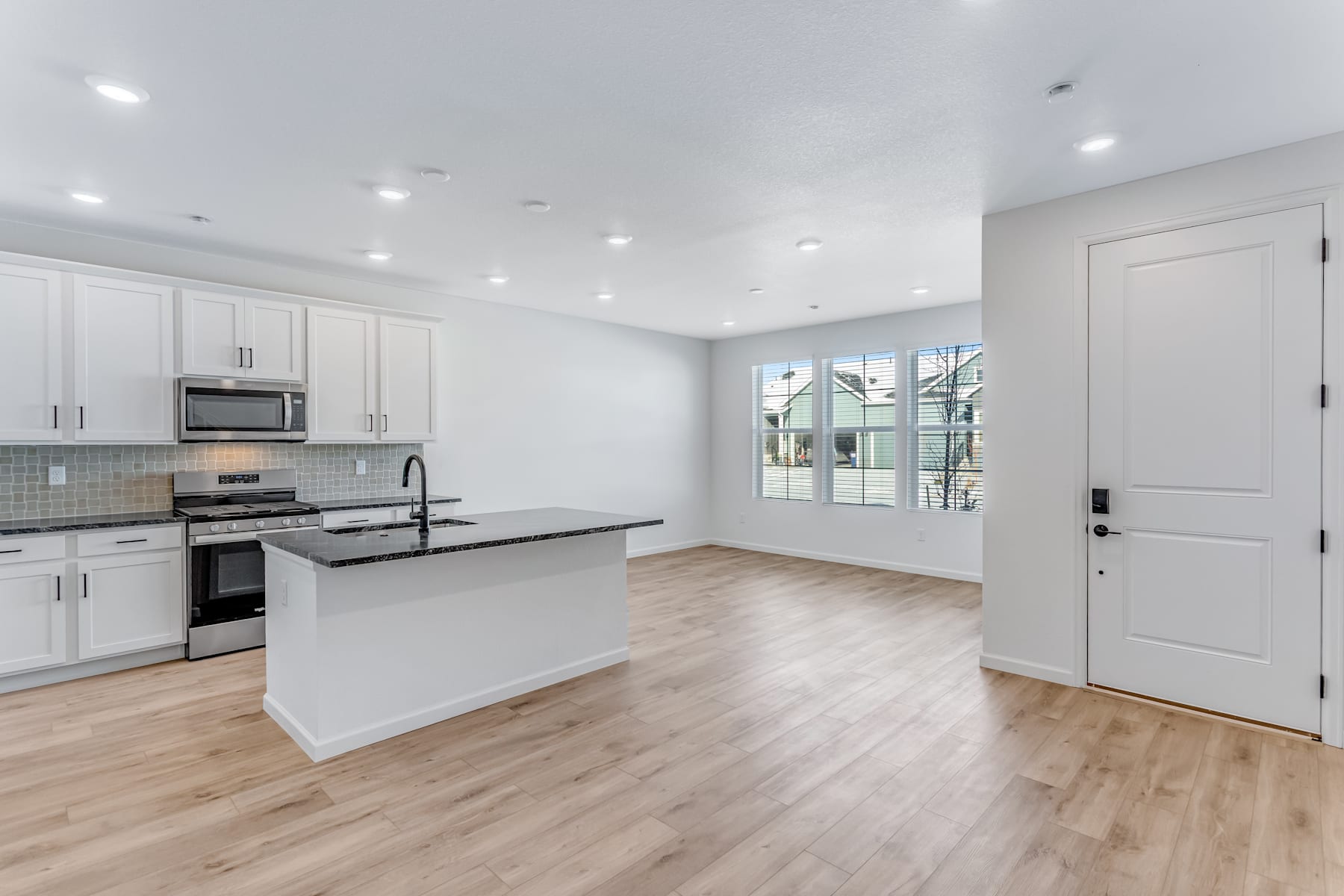 A modern, open-concept kitchen with white cabinets, stainless steel appliances, and hardwood floors, leading into a bright and airy living space with large windows.