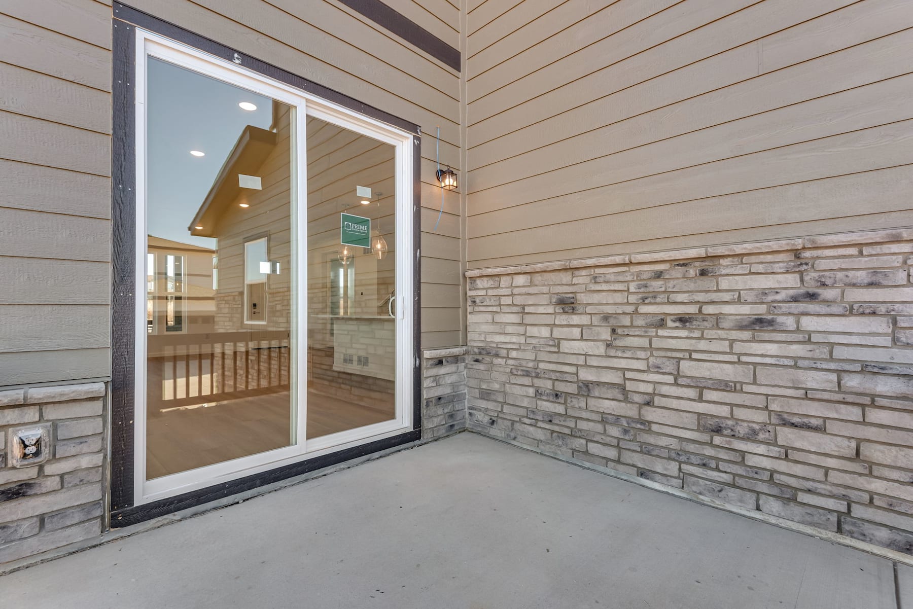 The image shows a patio or balcony area with a sliding glass door leading into what appears to be a residential interior. The foreground features a concrete floor, while the background showcases a stone or brick wall and wooden siding on the exterior of the building.
