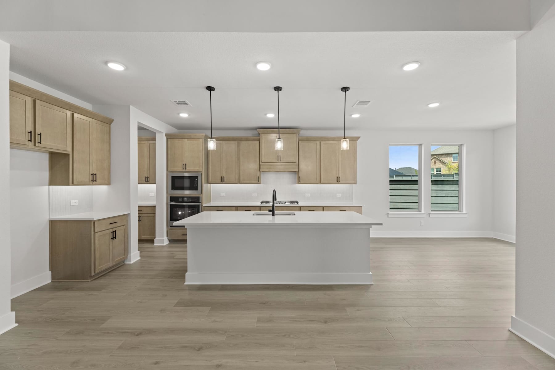 A modern, open-concept kitchen with light-colored wood cabinets, a large central island, and recessed lighting, set against a bright and airy background.