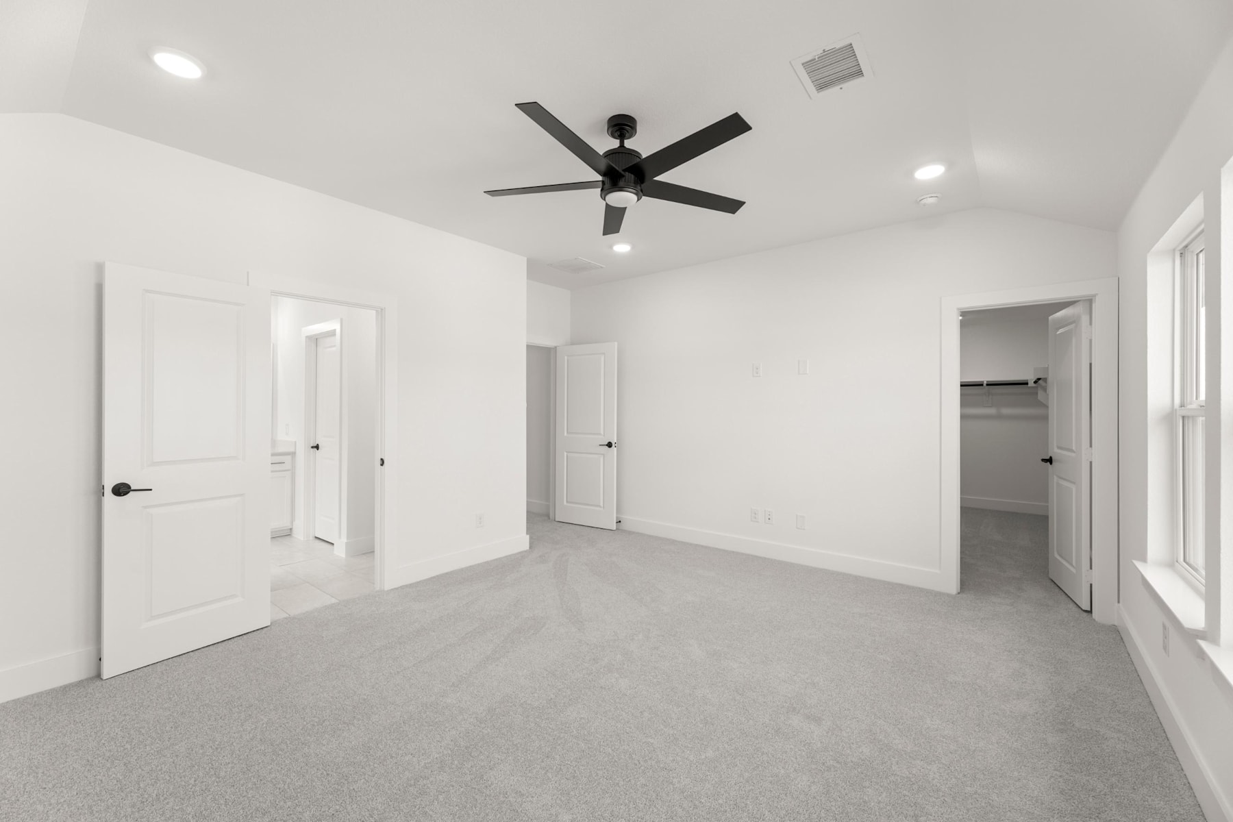 A spacious, minimalist room with a ceiling fan, white walls, and a gray carpeted floor.