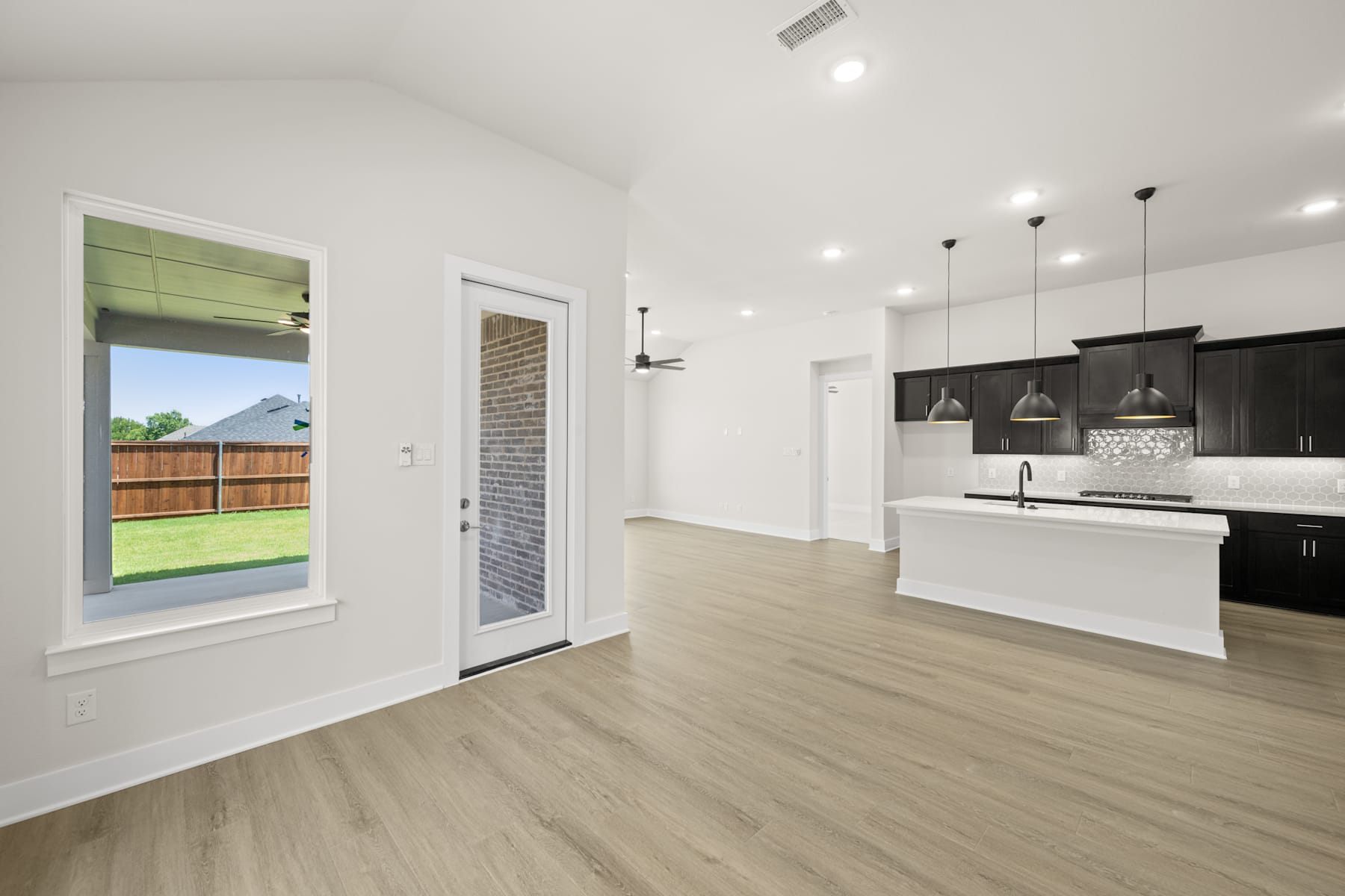 A modern, open-concept kitchen with white walls, dark cabinets, and a wooden floor leading to a backyard visible through a glass door.