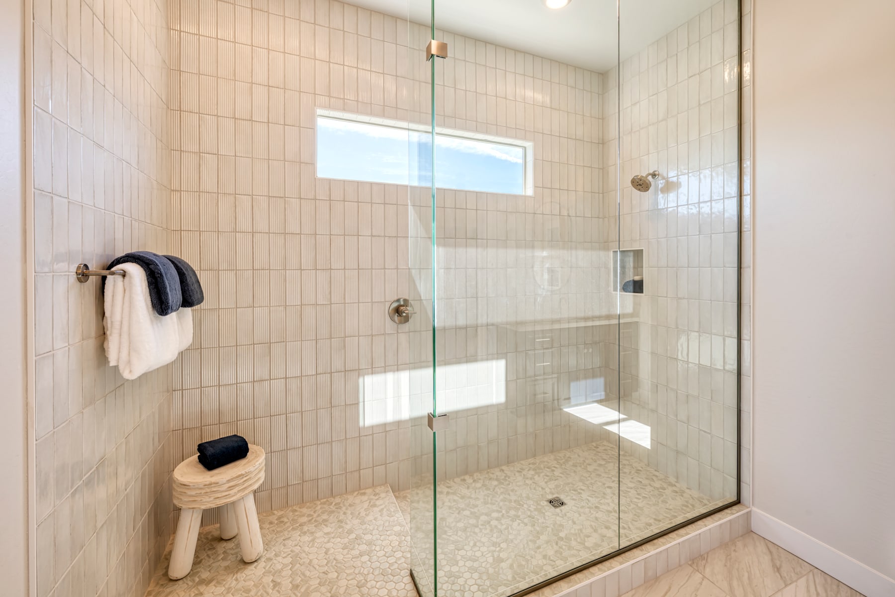 A modern, tiled bathroom with a glass shower enclosure, a wooden stool, and towels hanging on the wall.