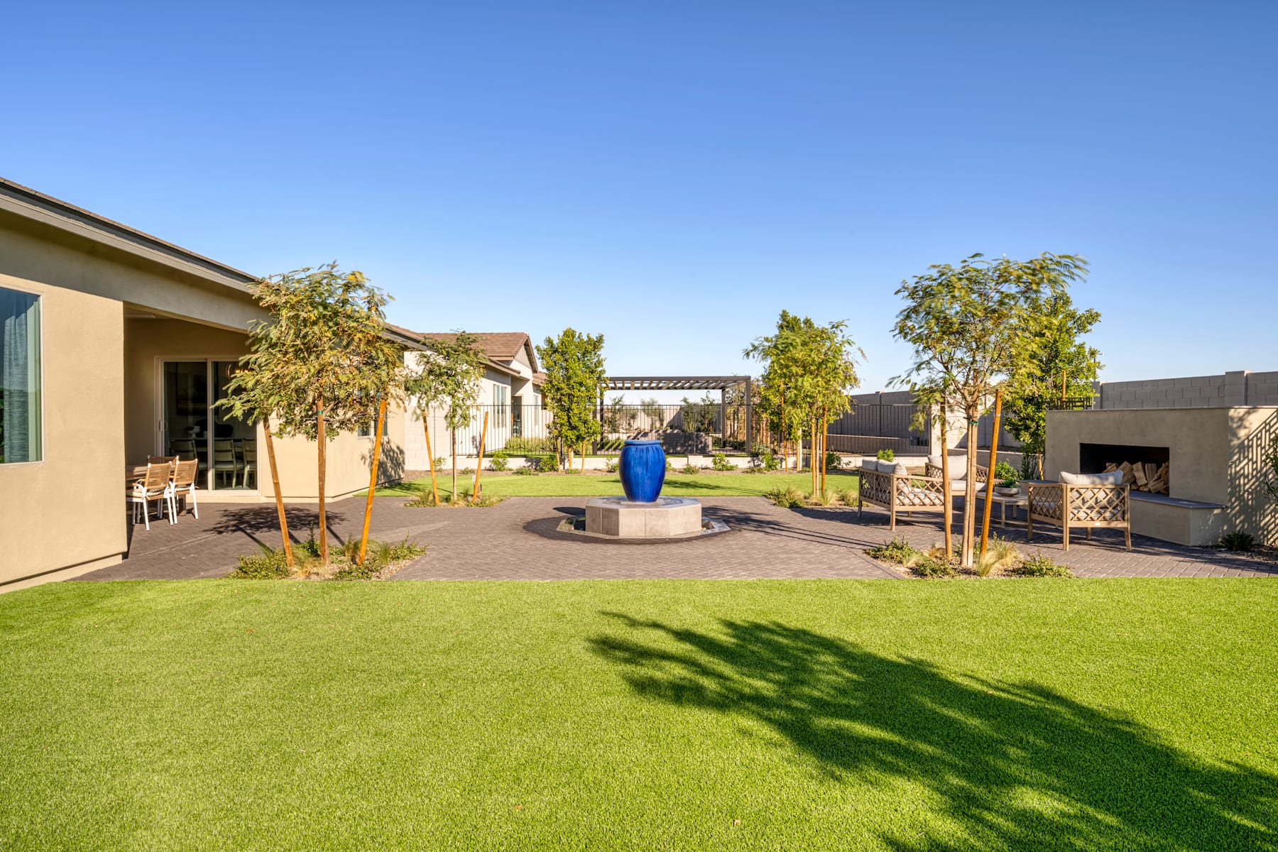 A well-manicured lawn with a decorative fountain in the center, surrounded by lush greenery and a modern building in the background under a clear blue sky.