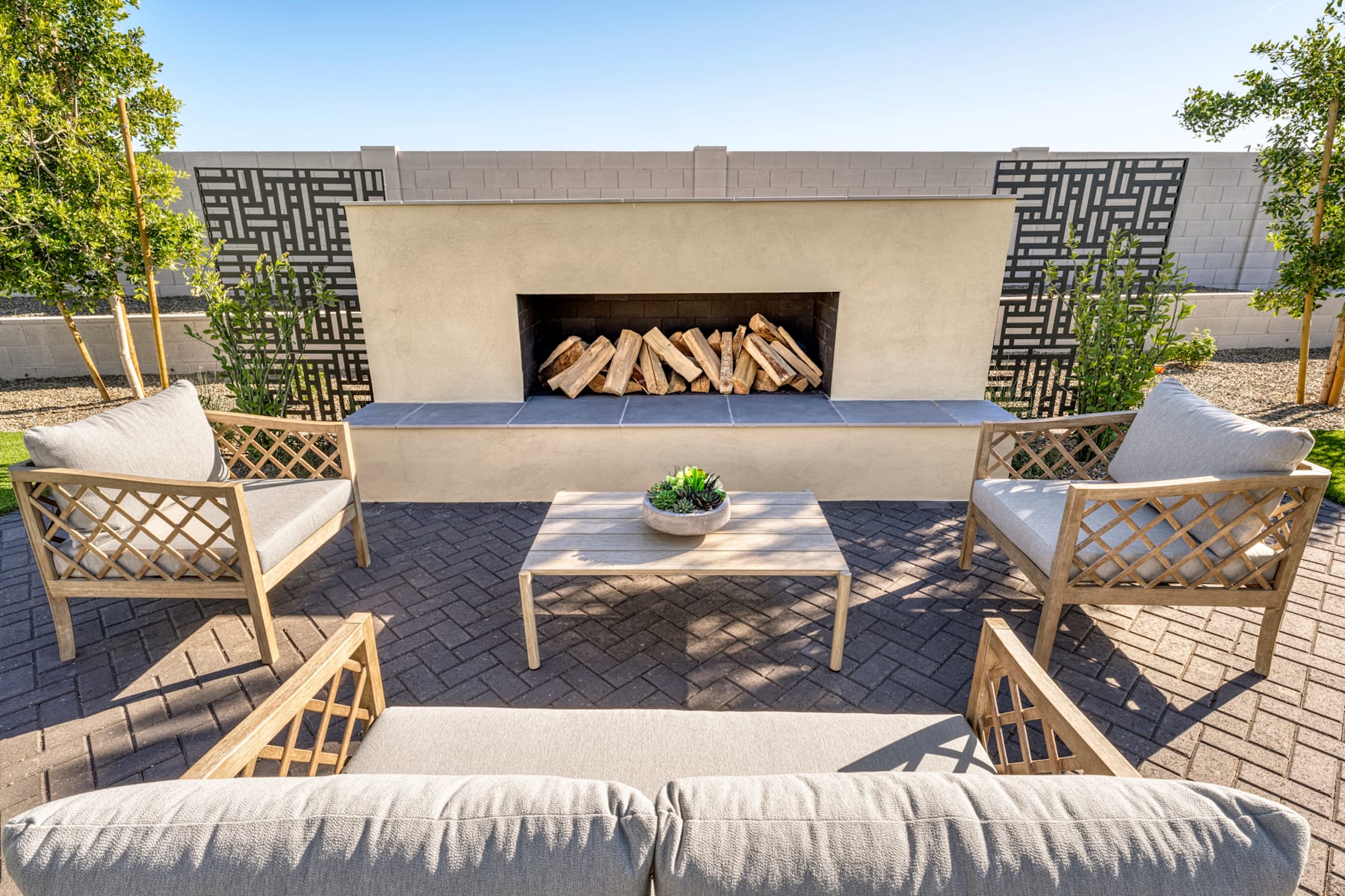 A cozy outdoor seating area with a fireplace, surrounded by lush greenery and a clear blue sky.
