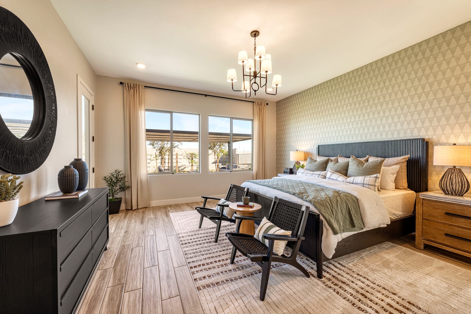 A cozy and well-appointed bedroom with a large bed, wooden furniture, and a chandelier overhead, set against a backdrop of patterned wallpaper and large windows.