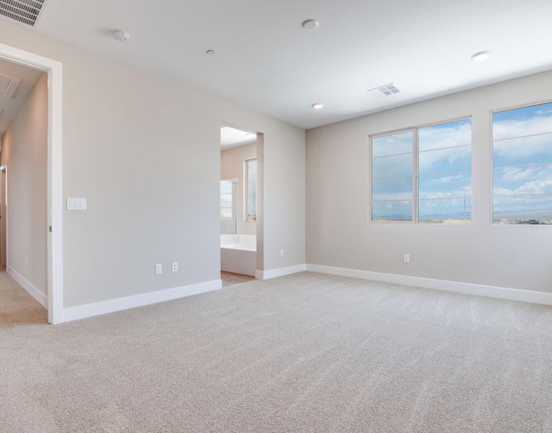 A spacious, well-lit room with white walls, a carpeted floor, and large windows offering a view of the sky.