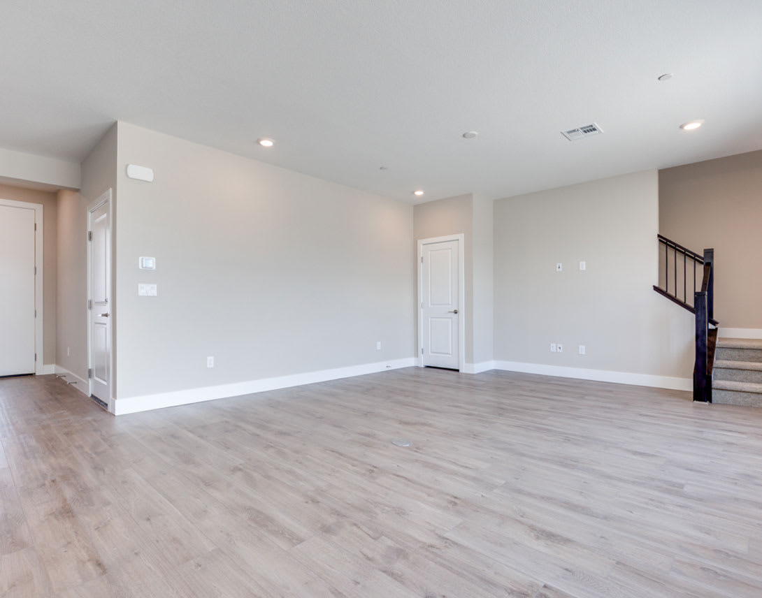 A spacious, well-lit room with hardwood floors, white walls, and a staircase visible in the background.