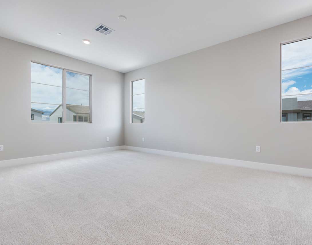 An empty, spacious room with white walls, a carpeted floor, and large windows providing natural light and a view of the exterior.