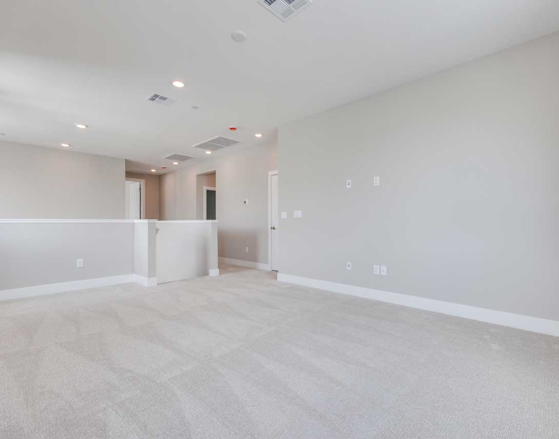 A spacious, well-lit room with white walls, a hardwood floor, and a partially visible hallway in the background.