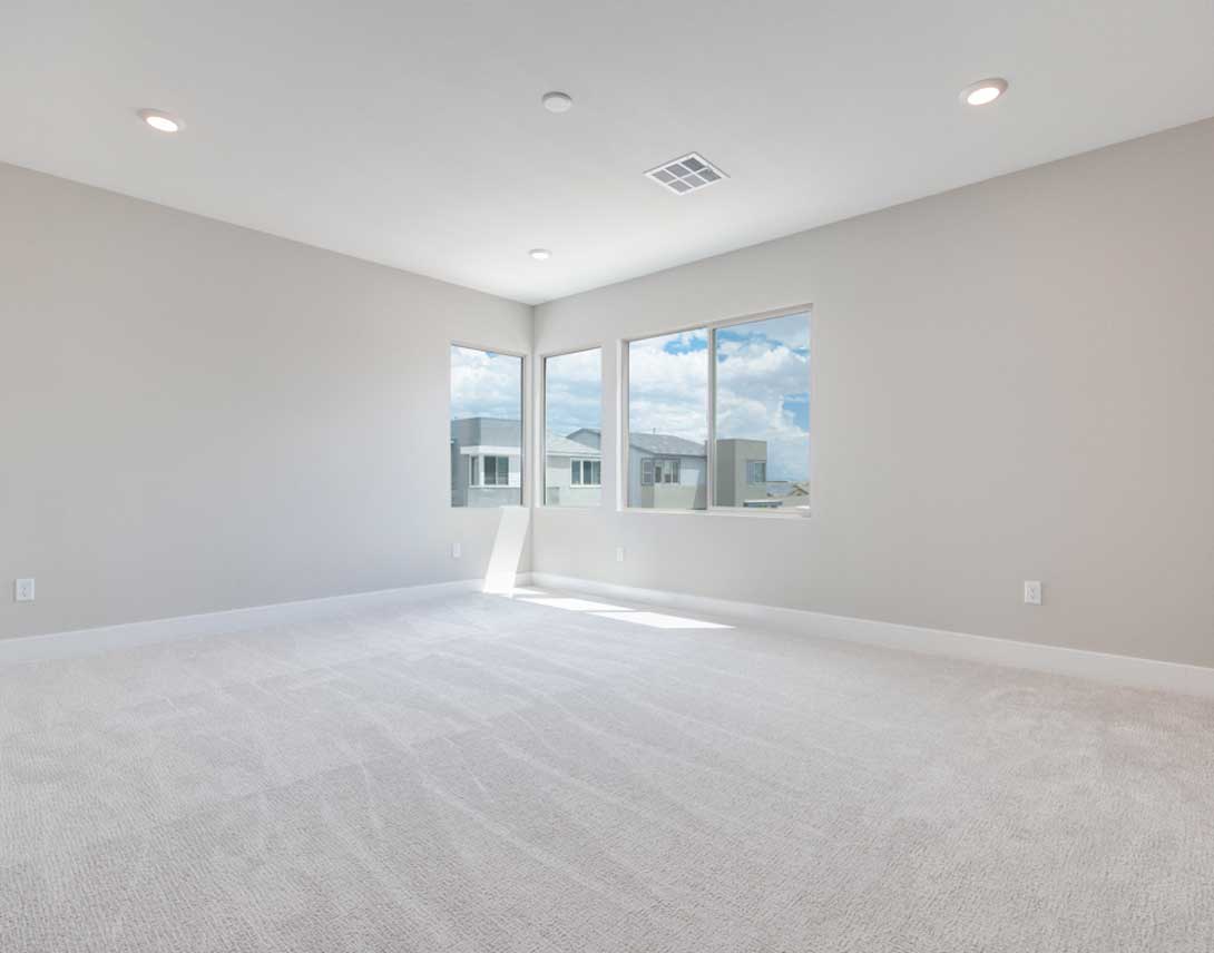 A spacious, well-lit room with white walls, hardwood floors, and large windows overlooking a residential neighborhood.