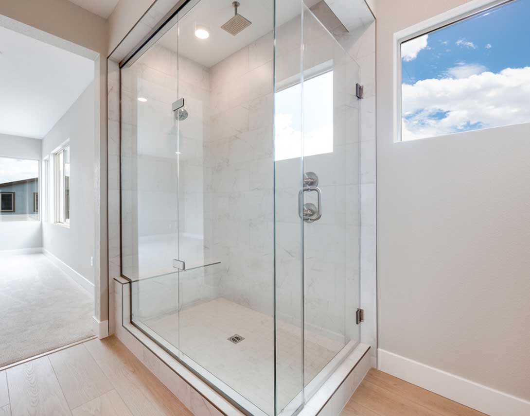 A modern, well-lit bathroom with a large glass shower enclosure, wooden flooring, and a view of the sky through the window.