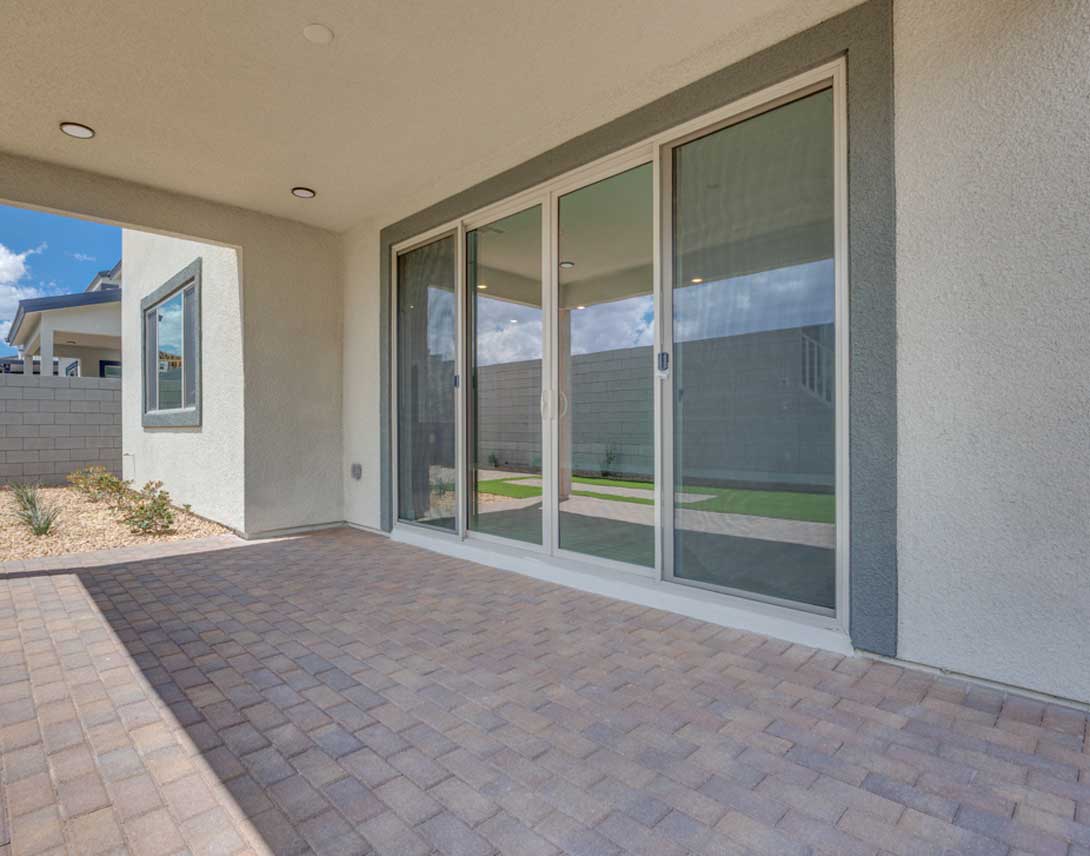 The image shows a paved patio area with sliding glass doors leading to a grassy backyard. The exterior walls of the building are painted in a neutral color scheme.