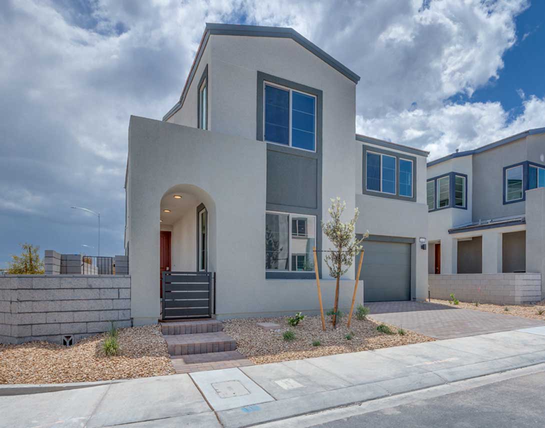 A modern, two-story residential building with a gray exterior, large windows, and a red front door, set against a backdrop of a cloudy sky and surrounded by a paved driveway and landscaping.
