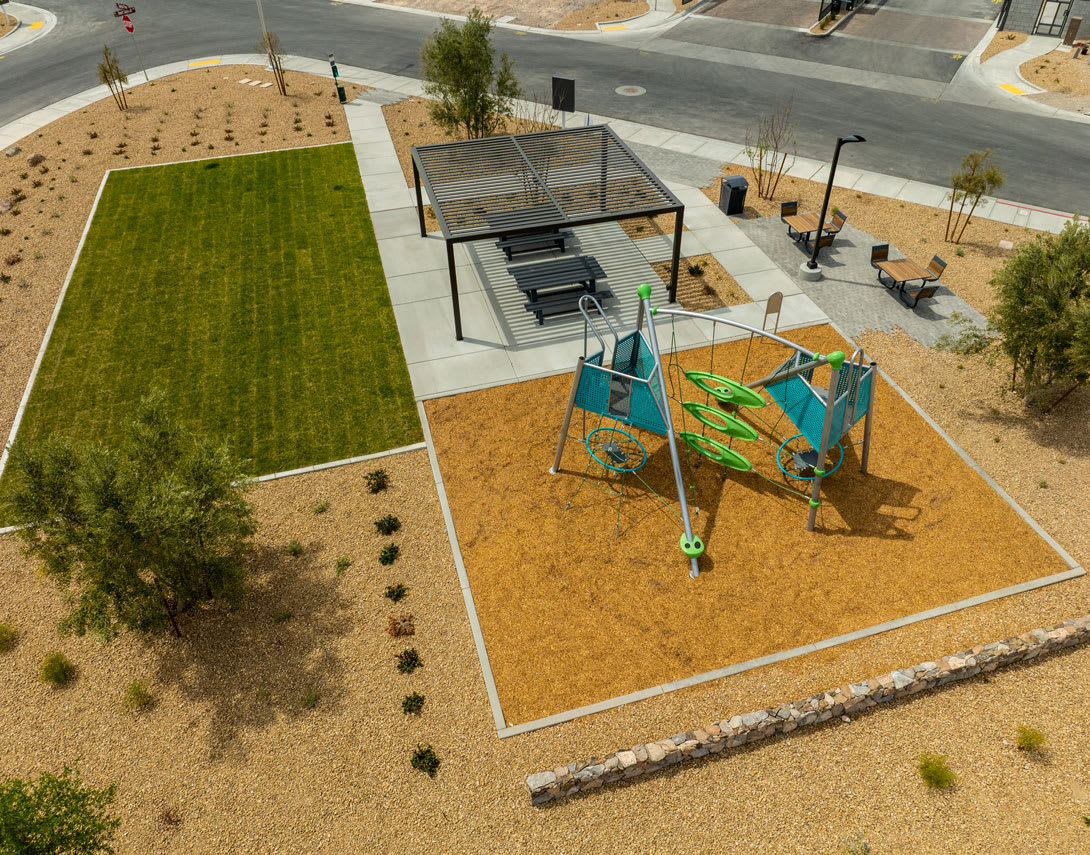 A vibrant outdoor public space with a grassy area, a colorful playground, and various seating options surrounded by paved walkways and landscaping.