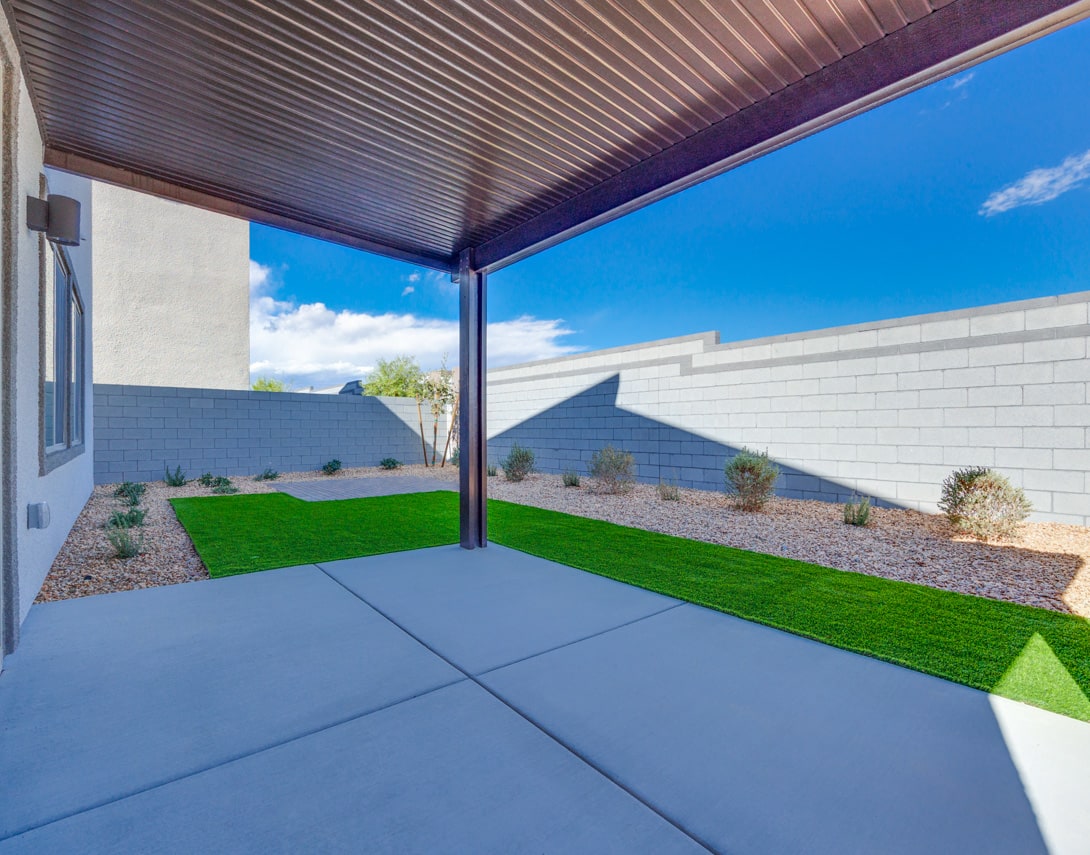 A covered patio with a concrete floor leads to a landscaped yard featuring a grassy area and desert-style plantings against a backdrop of a blue sky with clouds.