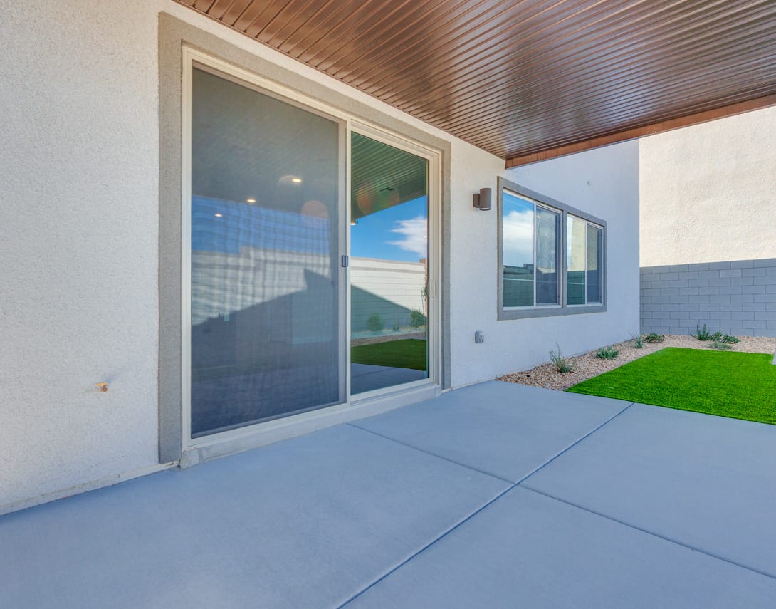 A modern patio with a concrete floor, wooden ceiling, and large sliding glass doors that offer a view of a grassy yard and distant mountains.