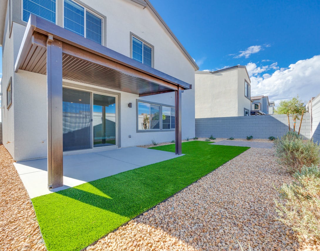 A modern, two-story residential building with a covered patio and a well-maintained yard featuring artificial grass and gravel landscaping.