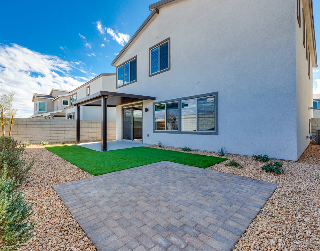 A modern, two-story residential building with a paved driveway, a covered patio, and a well-manicured lawn in the foreground, set against a clear blue sky with scattered clouds.