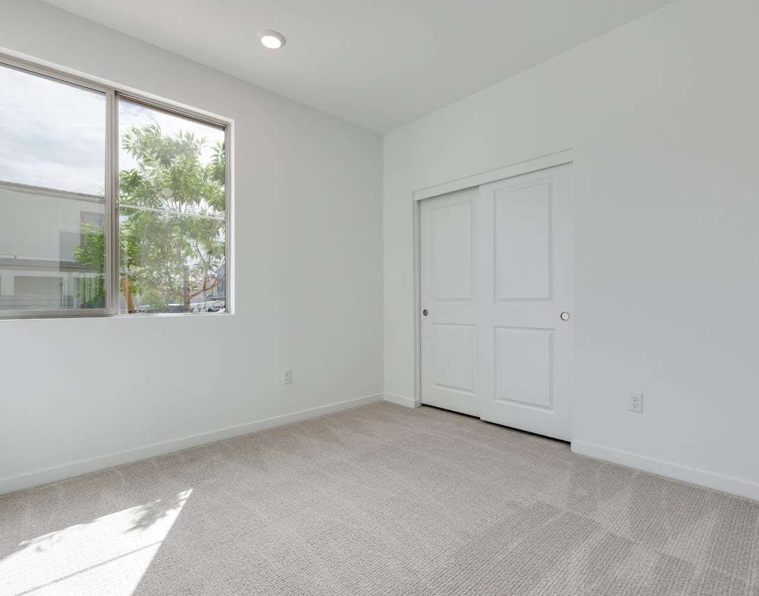 A bright, minimalist room with a large window overlooking greenery, a white door, and a light-colored hardwood floor.