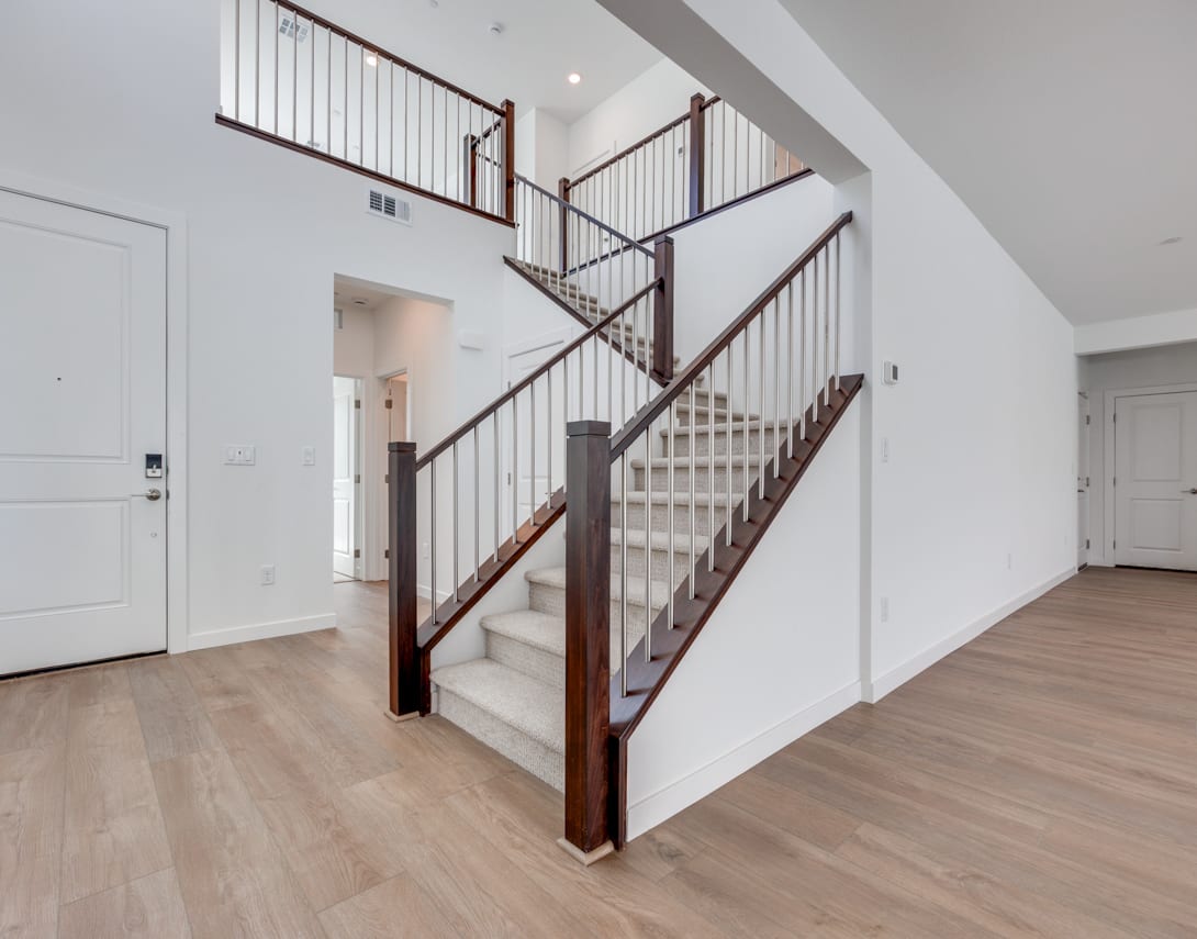 A modern, open-concept staircase with wooden steps and metal railings stands in a bright, airy space with white walls and hardwood floors.
