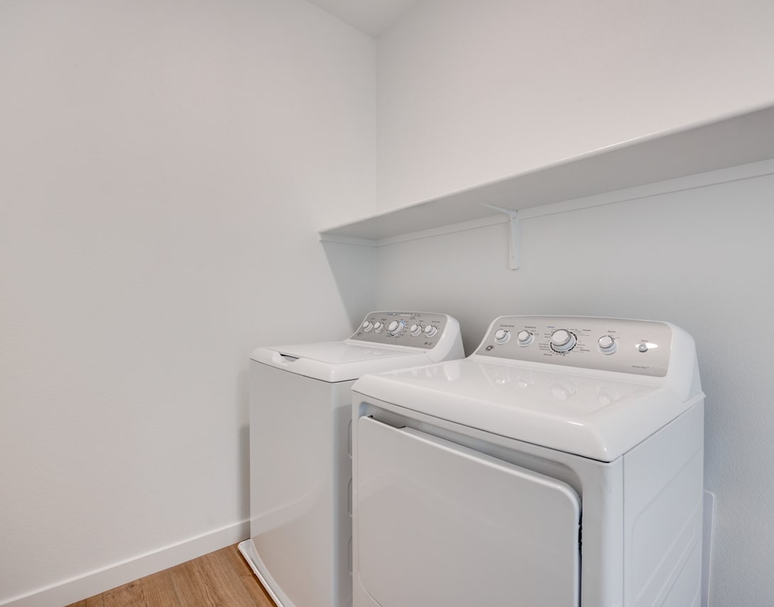 A modern, white washing machine and dryer set up in a clean, minimalist laundry room with bare walls and wooden flooring.
