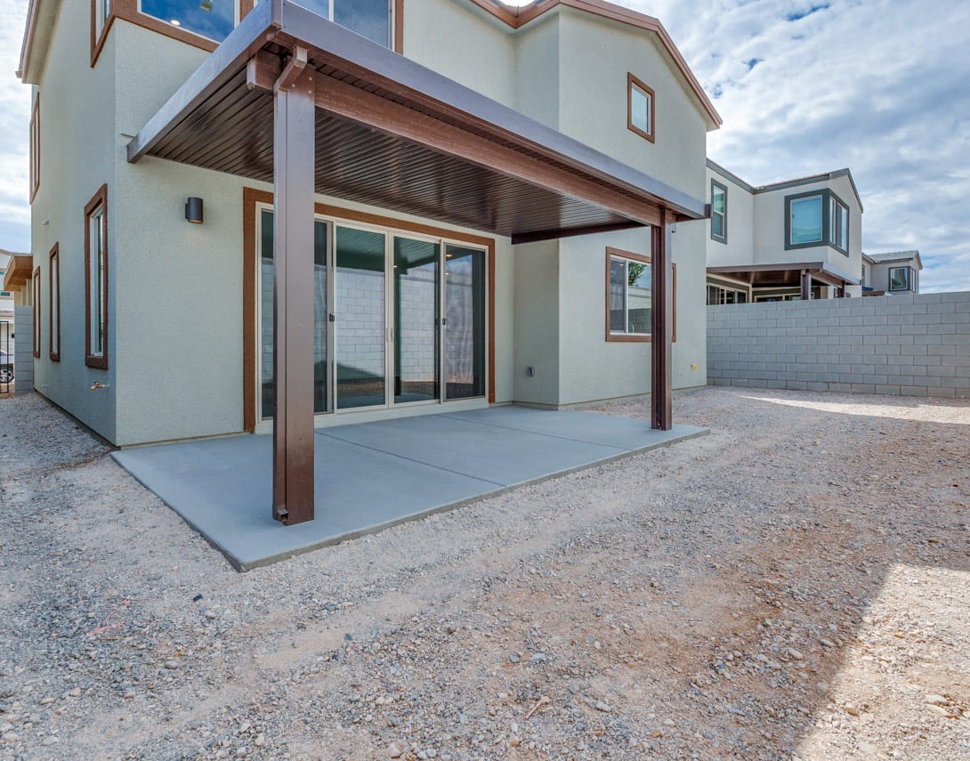 A modern, multi-story residential building with a covered patio area and a concrete patio in the foreground, set against a cloudy sky.