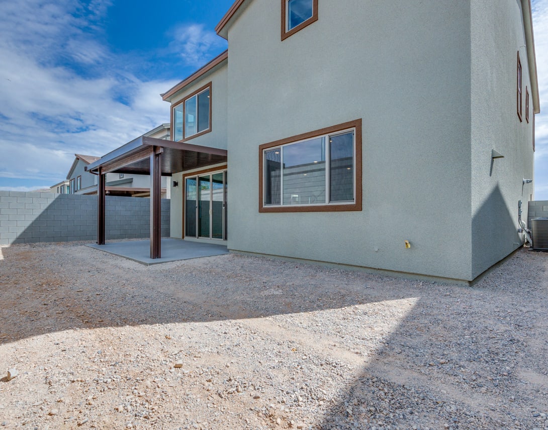 A modern two-story residential building with a covered patio and a gravel driveway in the foreground, set against a clear blue sky with scattered clouds.