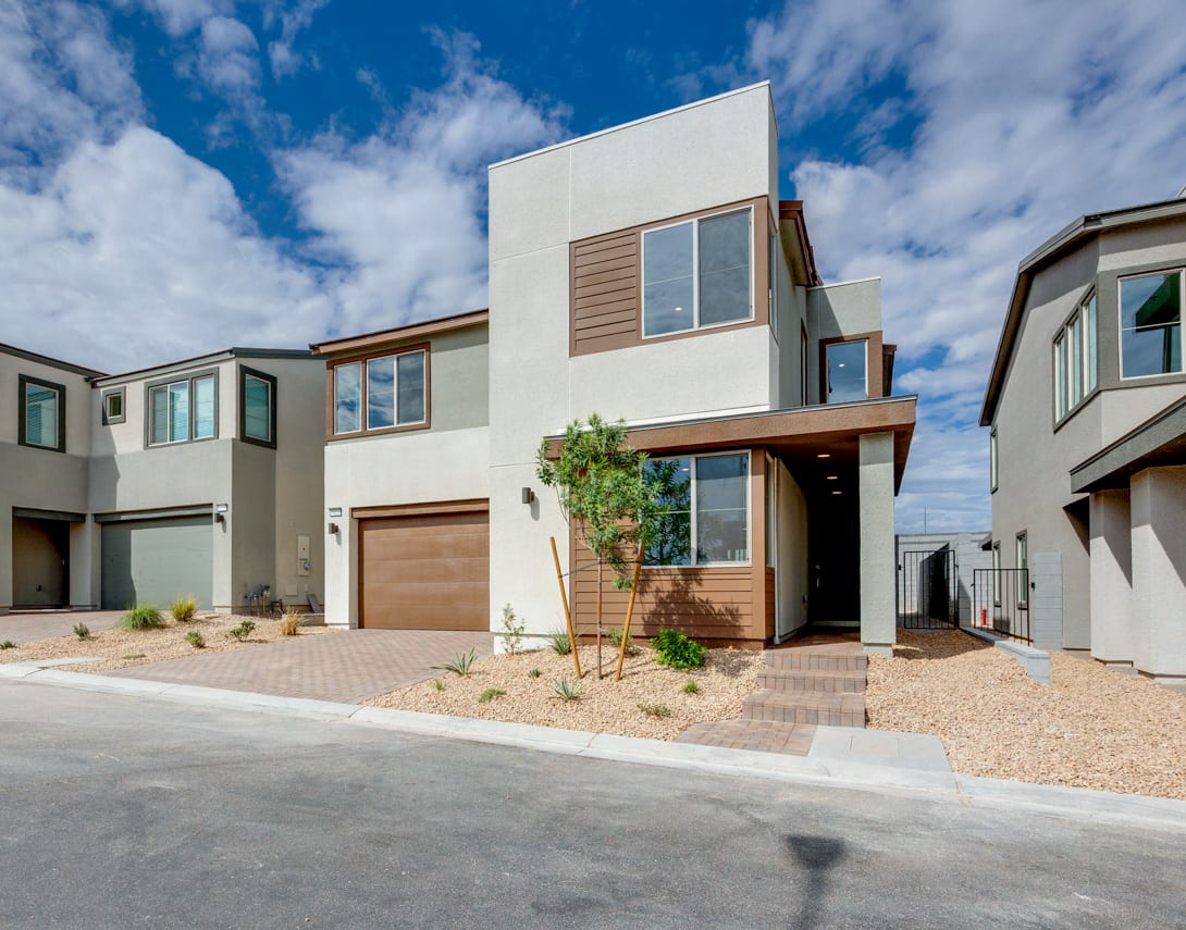 A modern, multi-story residential building with a garage, surrounded by a paved driveway and landscaped yard against a backdrop of a blue sky with scattered clouds.