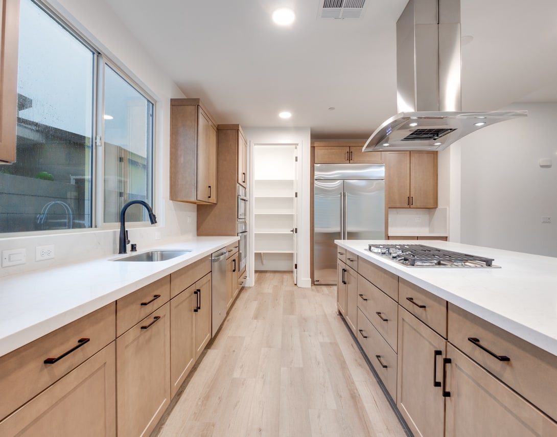 A modern, well-lit kitchen with light wood cabinets, white countertops, and stainless steel appliances, featuring a long, open layout with a window overlooking the exterior.