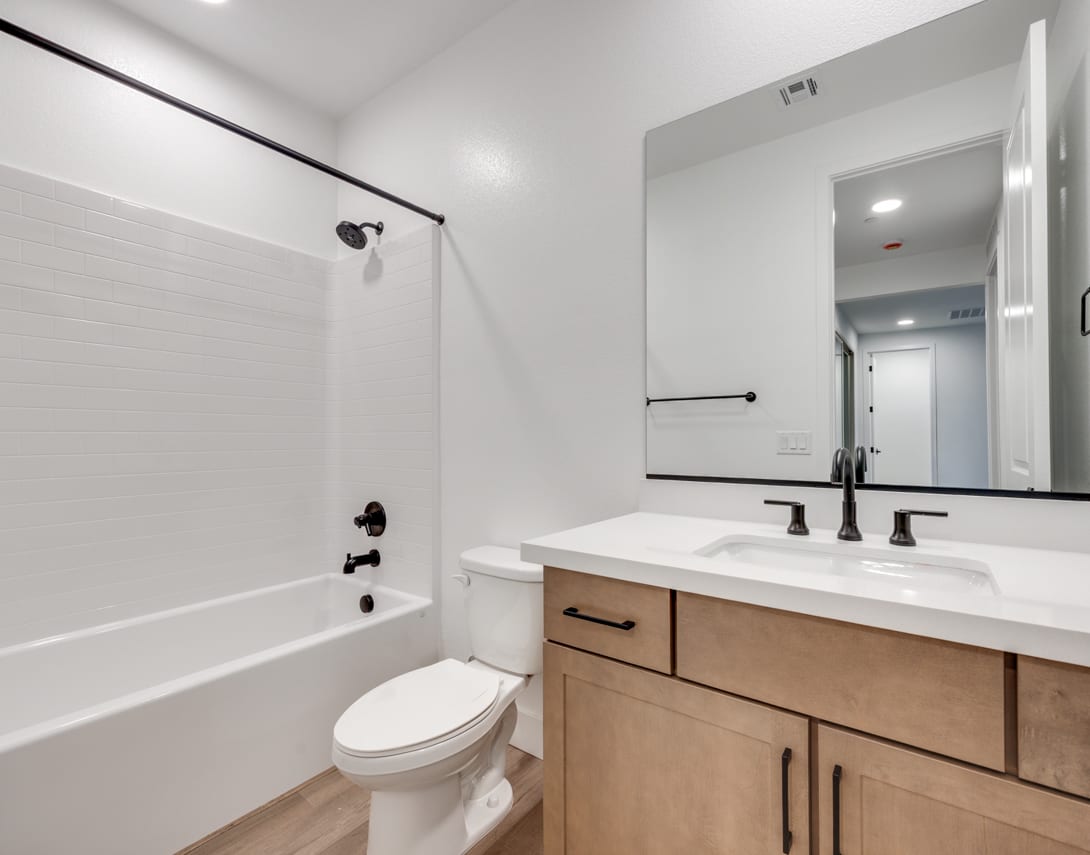 A modern, minimalist bathroom with a white vanity, a toilet, and a bathtub visible in the foreground, set against a neutral-toned wall and a doorway in the background.