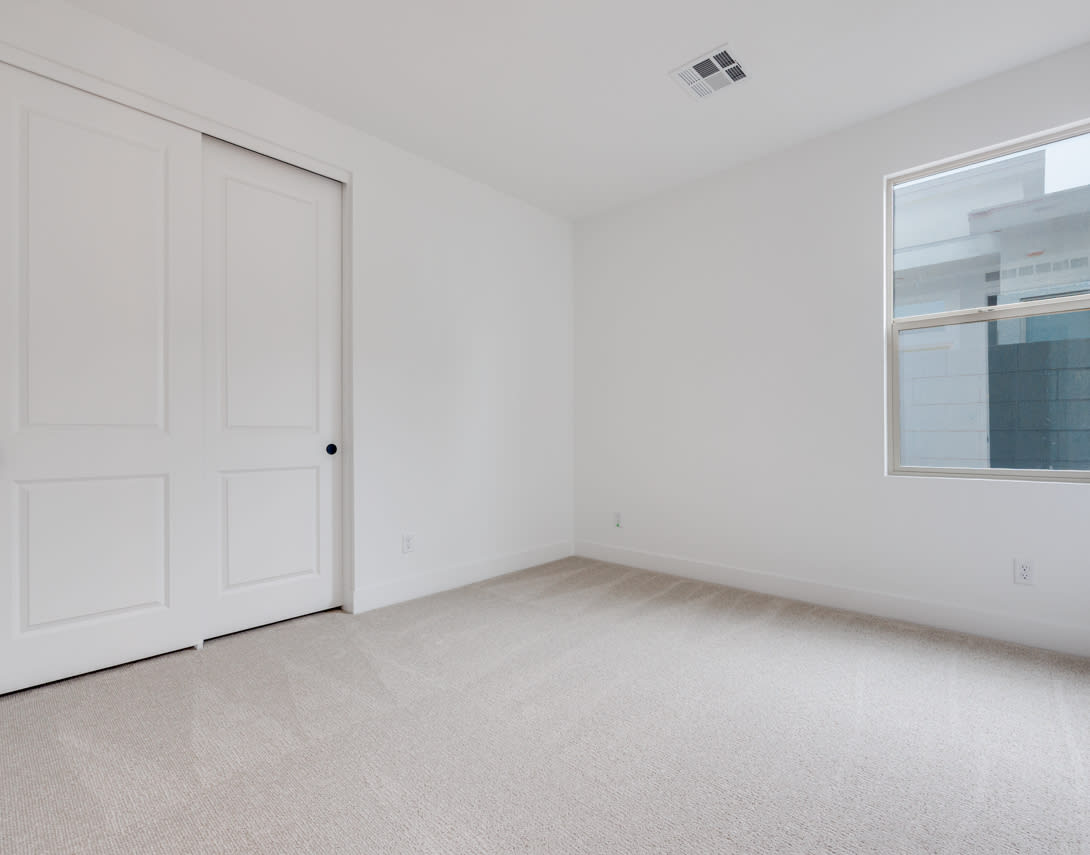 A simple, minimalist bedroom with a white wall, a closed door, and a window providing natural light.