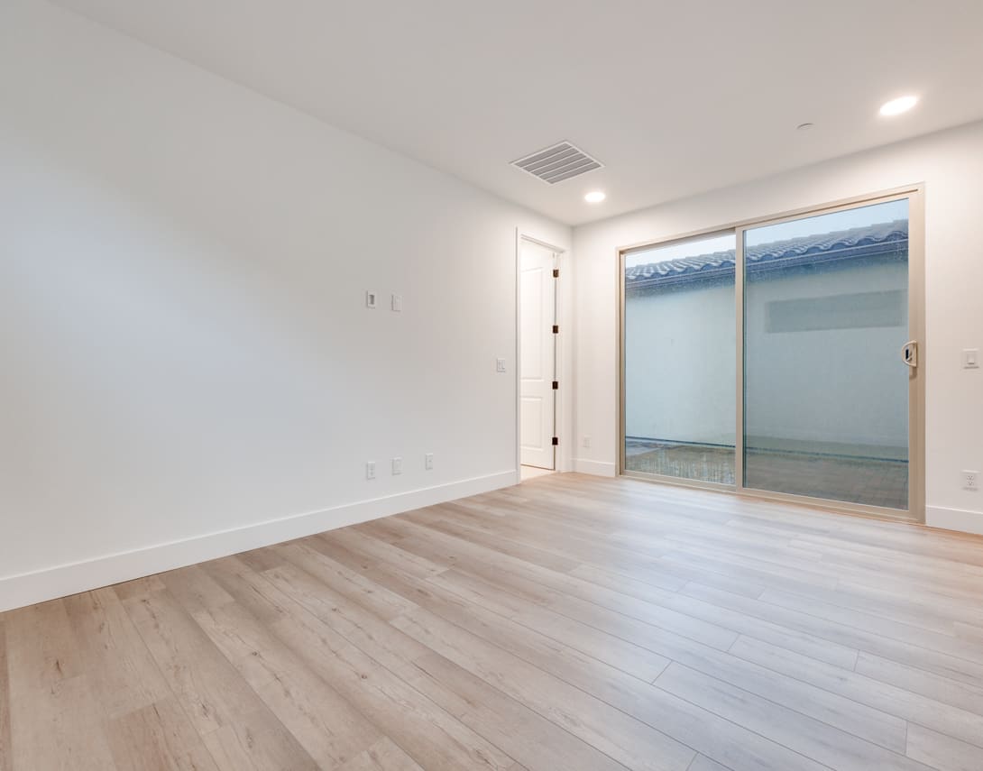 A spacious, well-lit room with a sliding glass door leading to an outdoor area, featuring a hardwood floor and a minimalist, clean design.