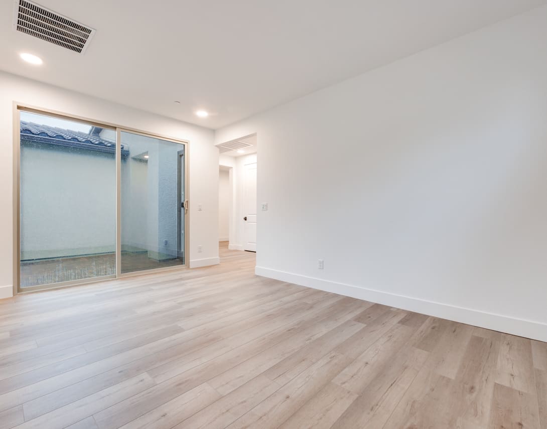 A spacious, well-lit room with clean, white walls and a wooden floor. A sliding glass door is visible in the background, providing a view of the exterior.