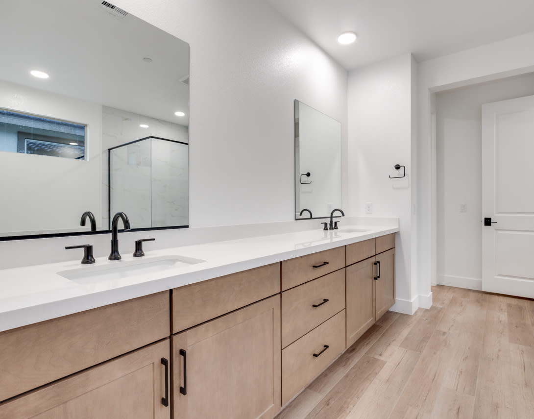 A modern and spacious bathroom with a double vanity, white countertops, and wooden cabinets, complemented by a glass shower enclosure and hardwood flooring.