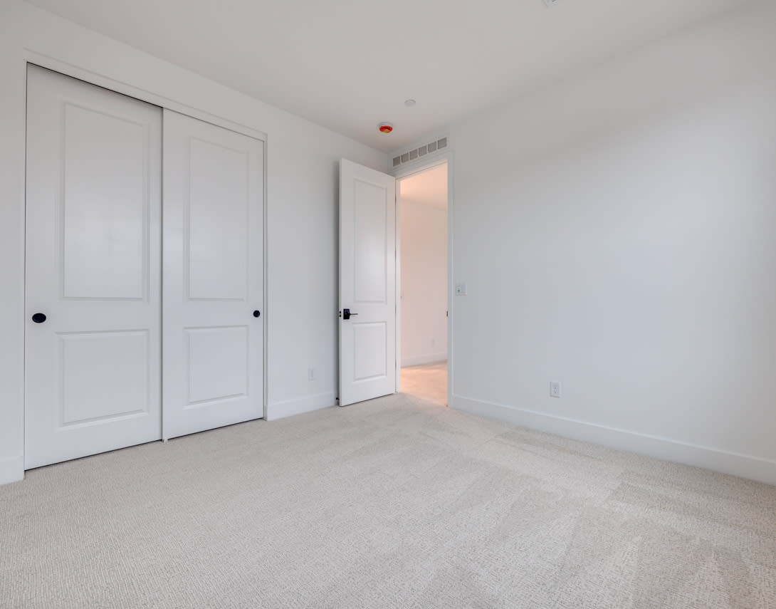 A spacious, minimalist bedroom with white walls, a carpeted floor, and two closed doors in the foreground.