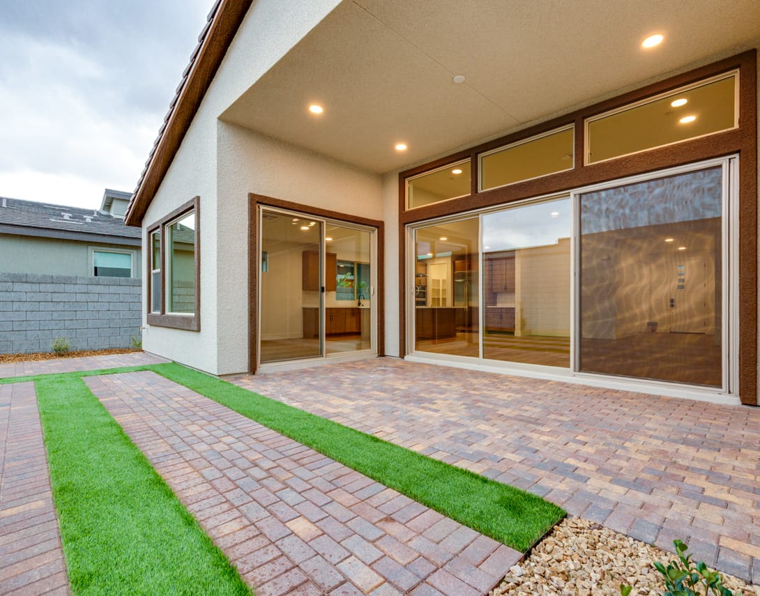 A modern, well-lit patio with a brick-paved floor, a lush green lawn, and large glass windows leading into the interior of the home.